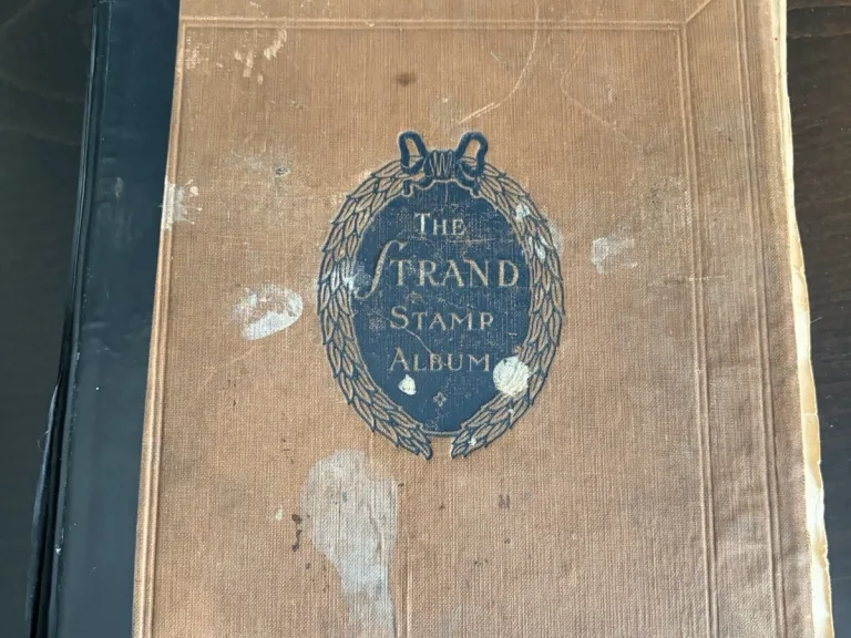 Worn tan cloth cover titled The Grand Stamp Album, with a dark blue spine and leaf-wreath device