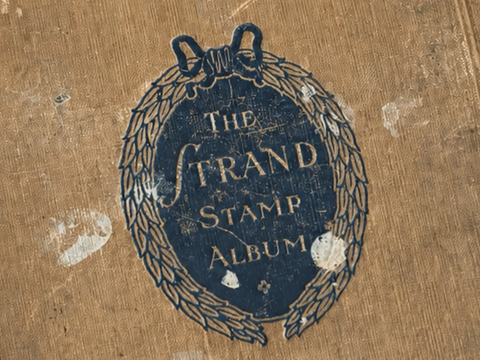 Close crop of the Grand Stamp Album’s original dark-blue leaf-wreath title device, tilted slightly against its worn tan cloth cover